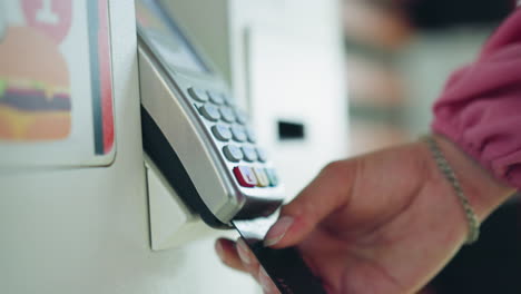 dama con camisa rosada, corto y cadena de muñeca inserta tarjeta negra en la máquina atm blanca para completar la transacción de pago mientras presiona el botón, fondo borroso con artículos poco claros