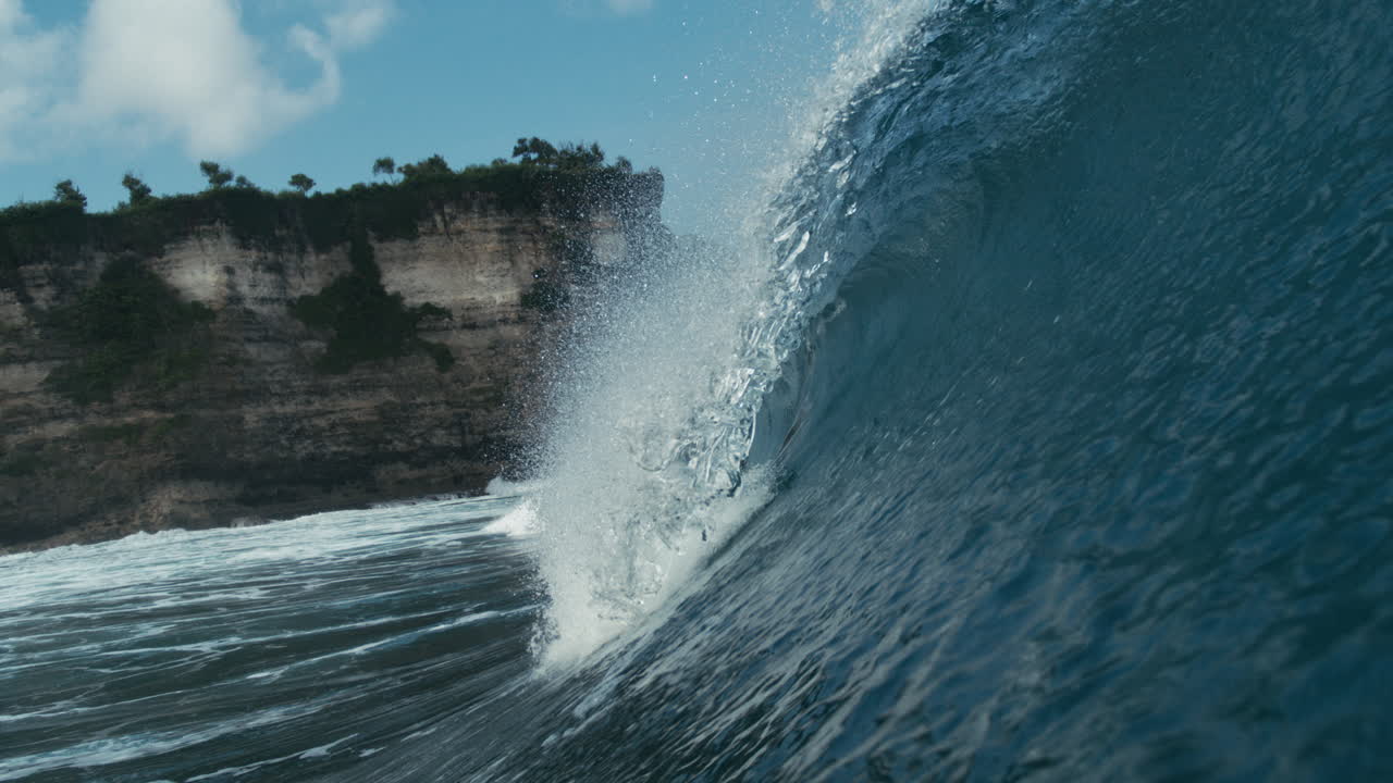 Curling tropical wave rolls past rocky point with tree lined shore visible, slow motion