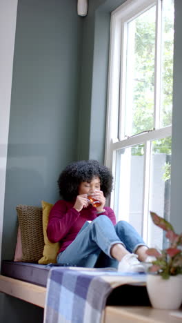 vertical video of biracial woman drinking tea and reading book at home, slow motion
