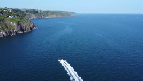 a following drone shot of a fast rigid inflatable boat that powers off to the right, whilst the camera tilts up revealing a beautiful coastal scene with stunning houses perched on the cliffs