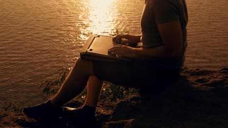 a man with a laptop in his hands is sitting on a cliff the background of the reflection of the sun path along the river at sunset.