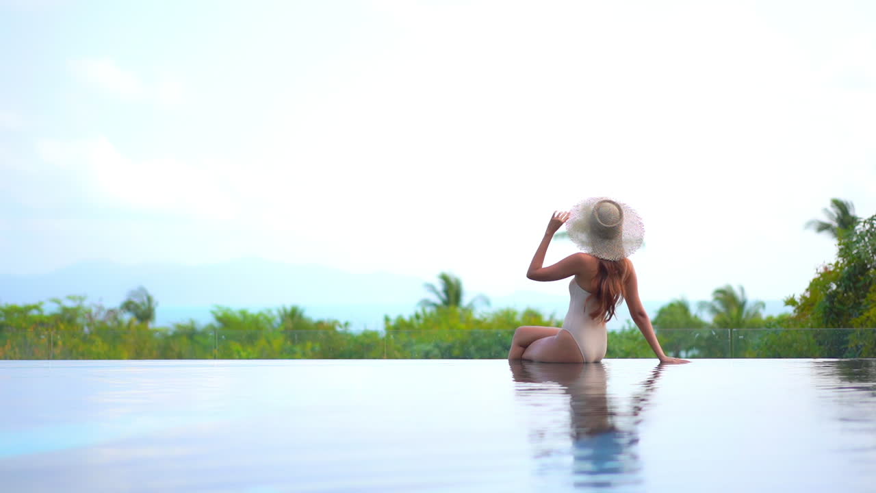 Model in Badeanzug und großem Hut sitzt am Rand des Infinity-Pools eines Luxusresorts auf einer tropischen Insel mit Blick auf eine üppige Landschaft und bewegt die Hand, um den Hut zu berühren.