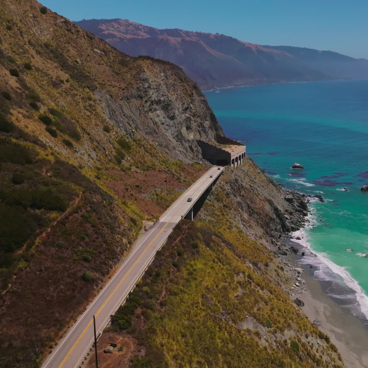 Two-lane highway with tunnels on the mountains of California coastline. Beautiful view of azure ocean at the shore. Top view