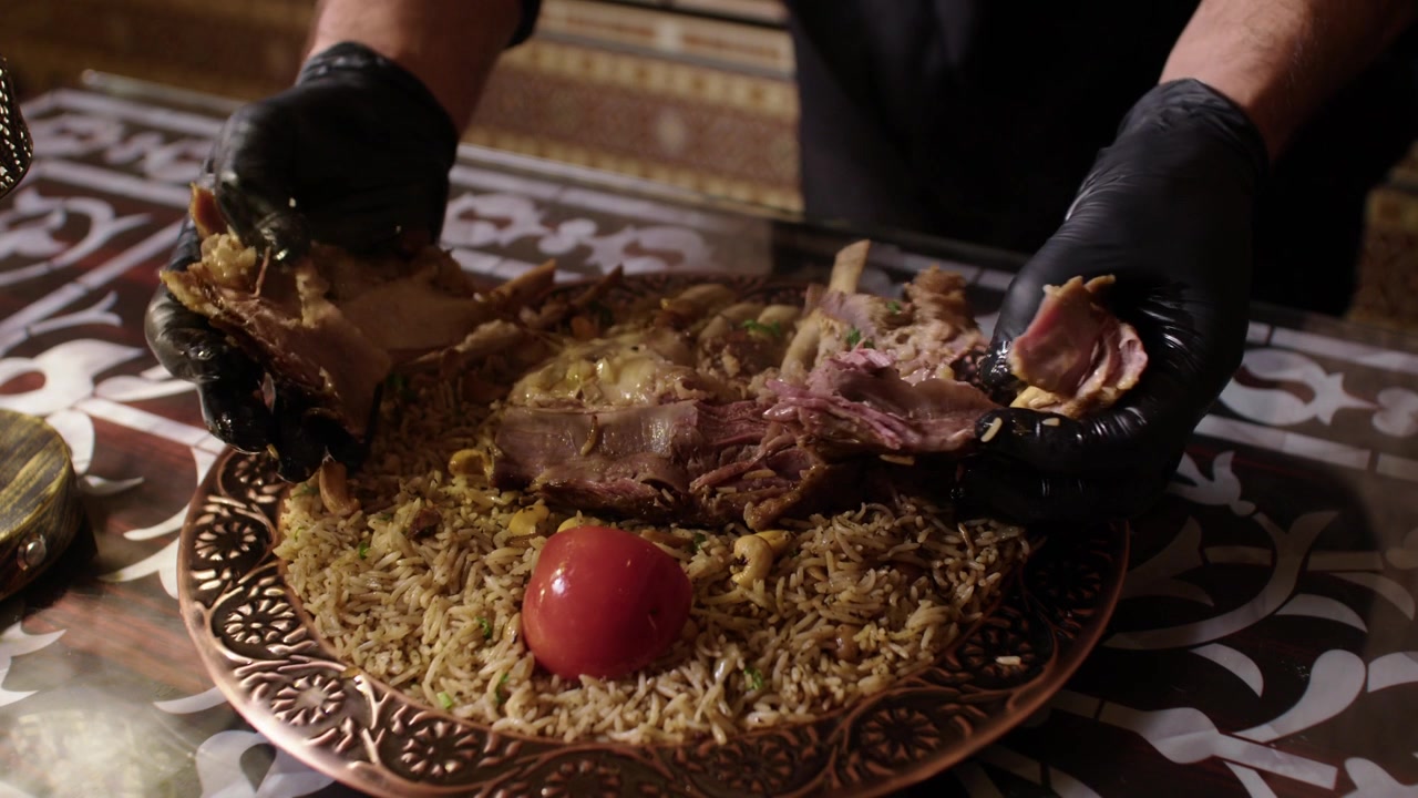 Chef in gloves serving slow-cooked meat over spiced rice on ornate plate indoors