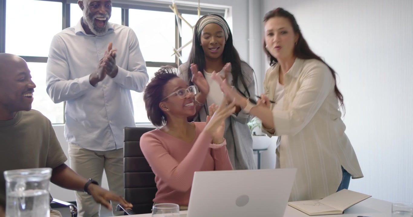 Diverse team in office host pointing charts sitting woman in pink sweater showing laptop clapping