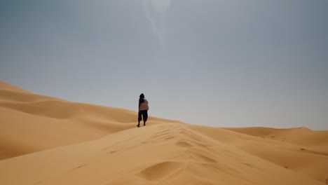 Person walks along the ridge of a golden sand dune in Merzouga, Sahara Desert, under clear midday skies