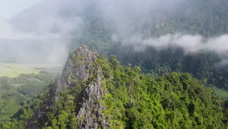 aerial flying past floating clouds with forested mountains of vang vieng with reveal of nam xay viewpoint