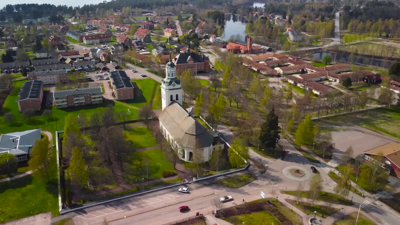Aerial drone foorage orbiting and spinning around a large white colored religious VImmerby church in a Swedish small rural gorgeous town with red rooftops and that has a lake on one side, sunny day.
