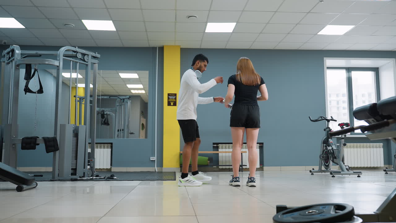trainer hands dumbbell to woman inside bright spacious gym as he supervises her preparation for knee lunge workout, emphasizing guidance, support, and strength training