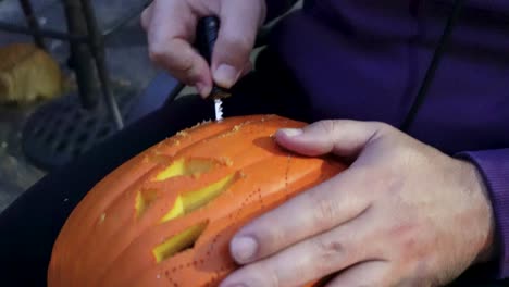 primer plano de hombre vestido con capucha tallando cuidadosamente una calabaza naranja en halloween