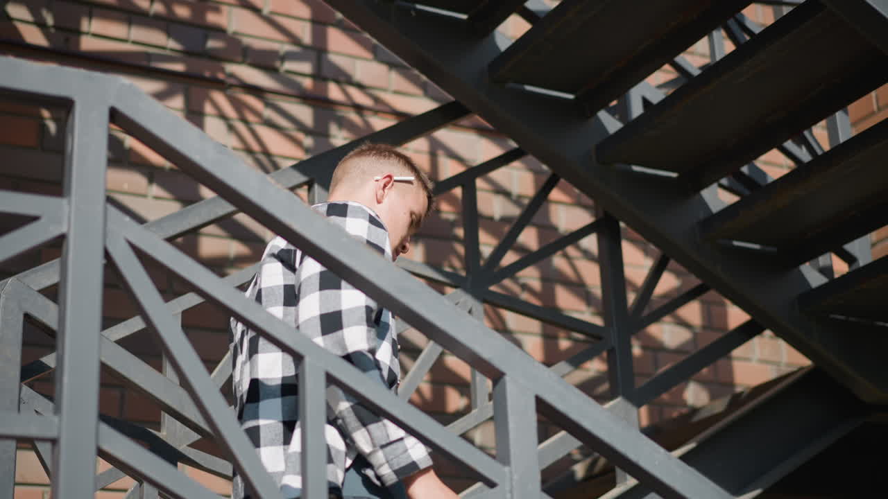 side view of boy descending outdoor metal balcony staircase against brick wall removes cigarette from ear raises hand to lighter sparks cigarette tip inhales smoke with sunlight reflecting off face
