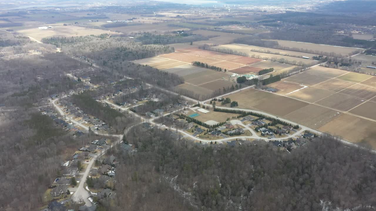 Vista aerea ad alta quota di un vasto paesaggio con un quartiere residenziale periferico immerso in spogli boschi invernali, adiacente a vasti campi agricoli sotto un cielo parzialmente nuvoloso