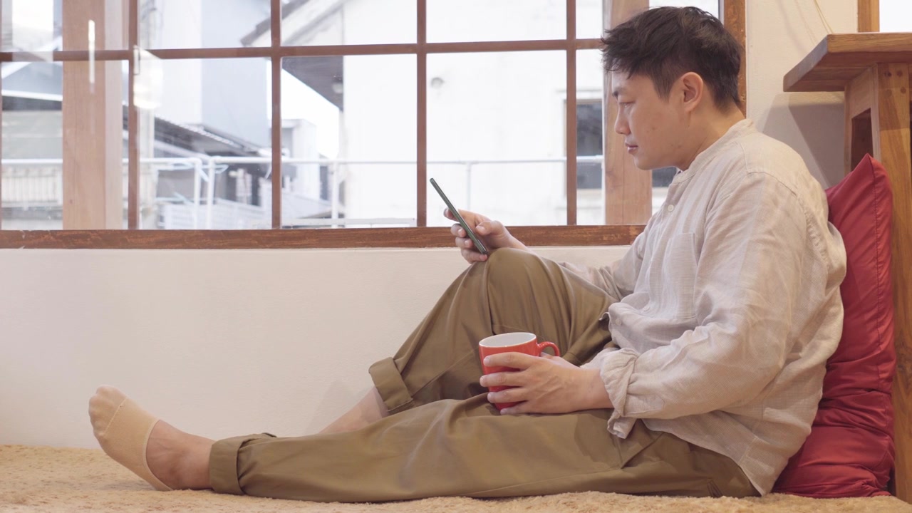 自宅の窓際で赤いコーヒーマグを手に持ち、スマートフォンを操作する男性