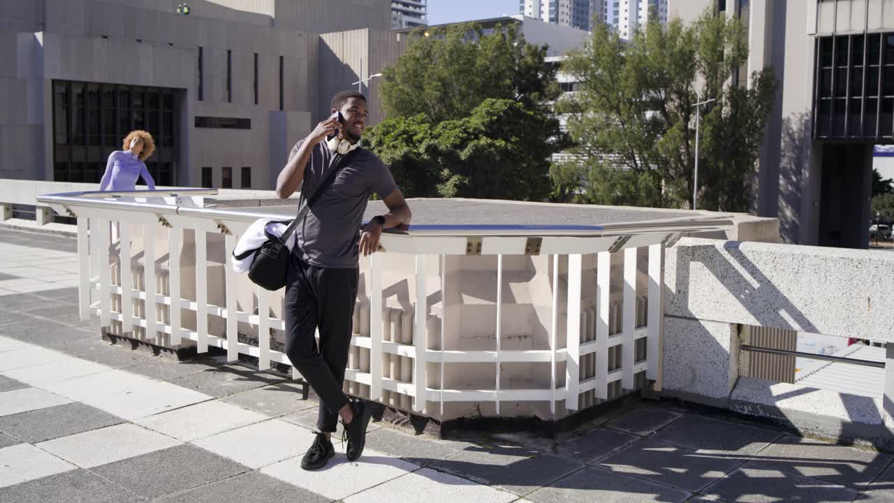 African American man with duffel walking rooftop plaza, checking phone after ping, answering call