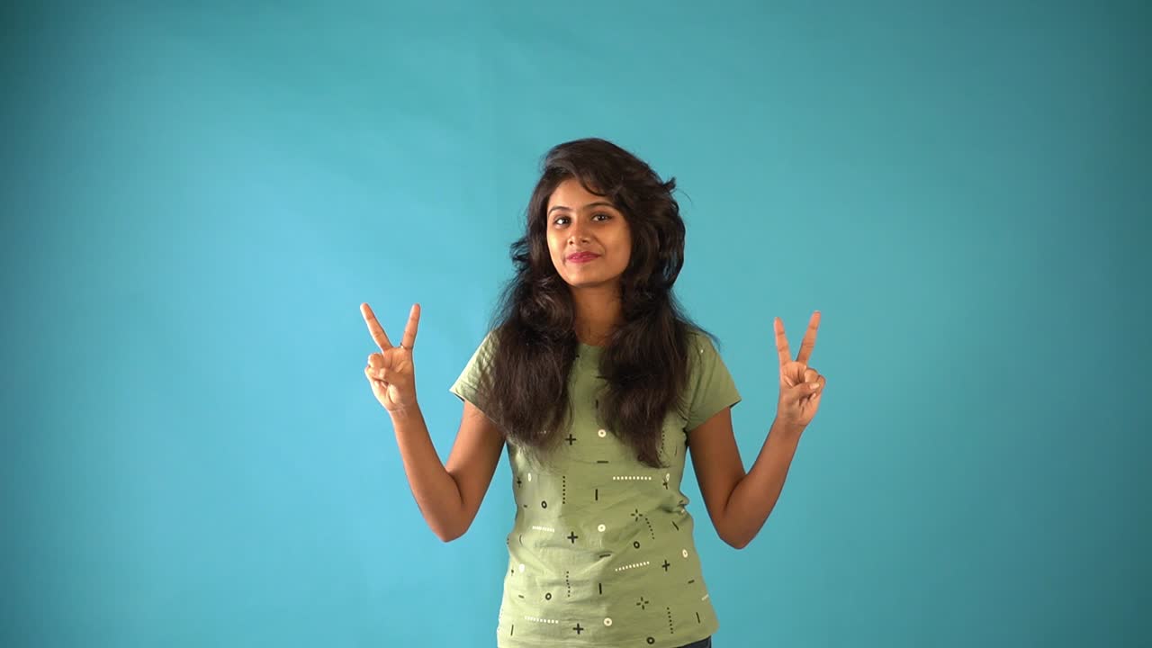 A young Indian girl in green t-shirt showing the symbol of peace looking at the camera standing in an isolated blue background studio