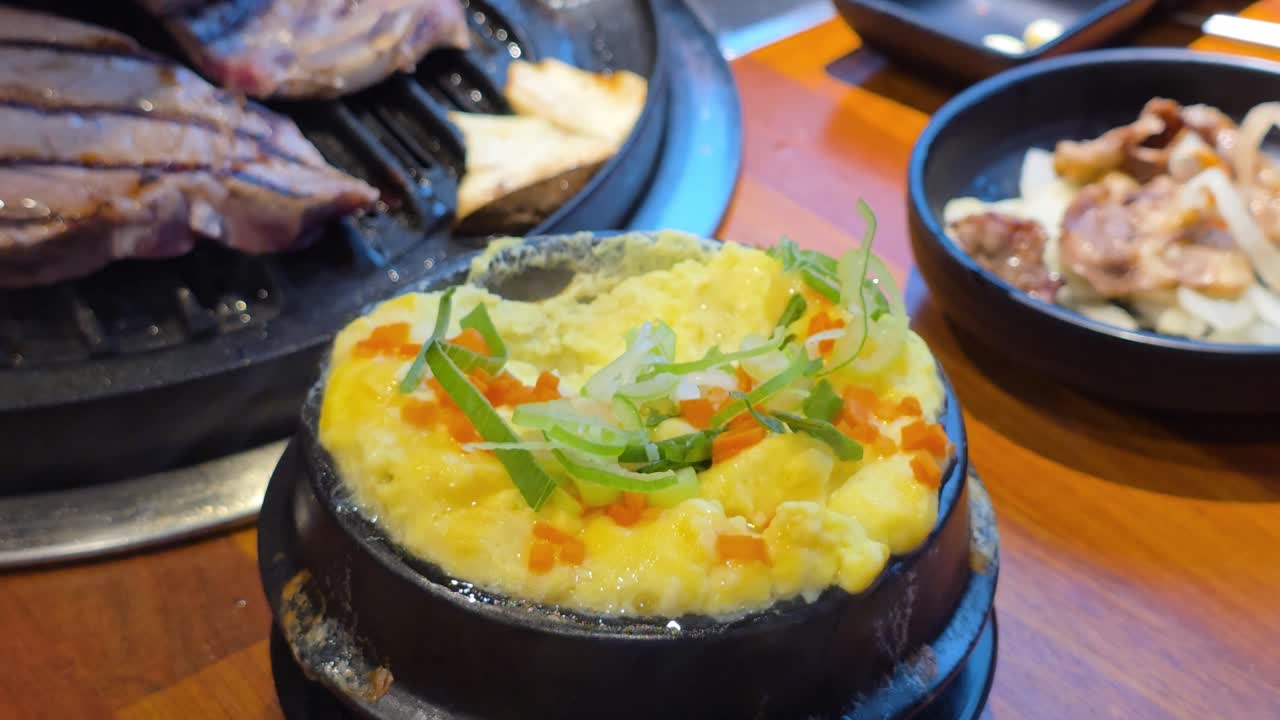 A close-up showcases a savory Korean steamed egg dish (Gyeran Jjim) in a black earthenware bowl, garnished with chopped scallions and carrots, at a bustling Korean BBQ restaurant in Seoul