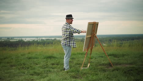 a craftsperson, wearing a black hat, checkered shirt, and jeans, is energetically painting a large canvas in the middle of a peaceful grass field