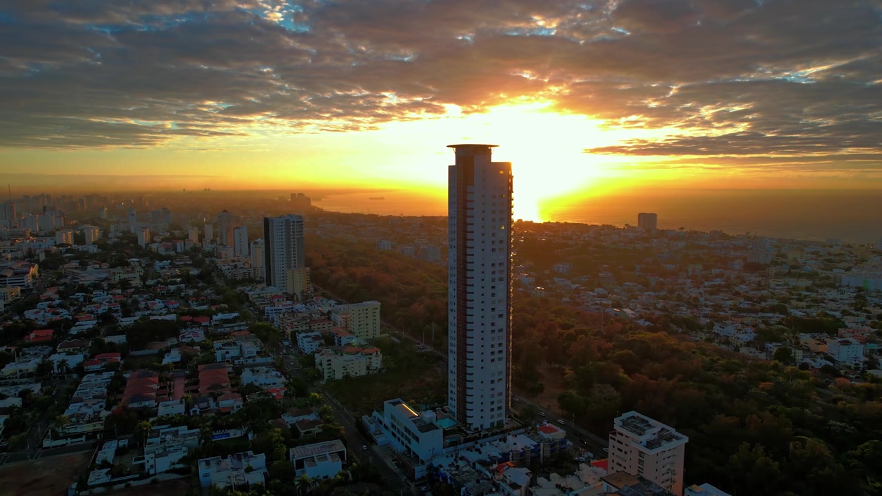 Tomada aérea de drone em ângulo alto avançando e descendo em direção ao arranha-céu Caney Tower, capturando o sol dourado brilhante se pondo atrás do edifício sobre o mar.