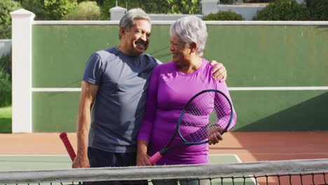 video de una feliz pareja de adultos mayores de dos razas sosteniendo raquetas en la cancha de tenis