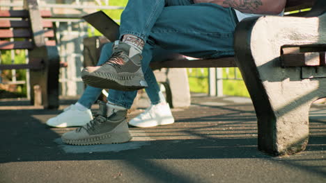 close-up of legs of two people seated outdoors on wooden benches, wearing casual sneakers and jeans, with sunlight casting shadows and greenery visible in the background