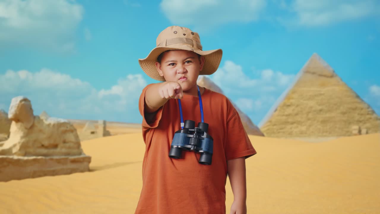 Asian Boy With A Hat And Binoculars Pointing His Finger Towards You And Furiously Speaking Against Blaming You While Traveling In Giza Pyramid. Boy Researcher, Travel Tourism Adventure Concept