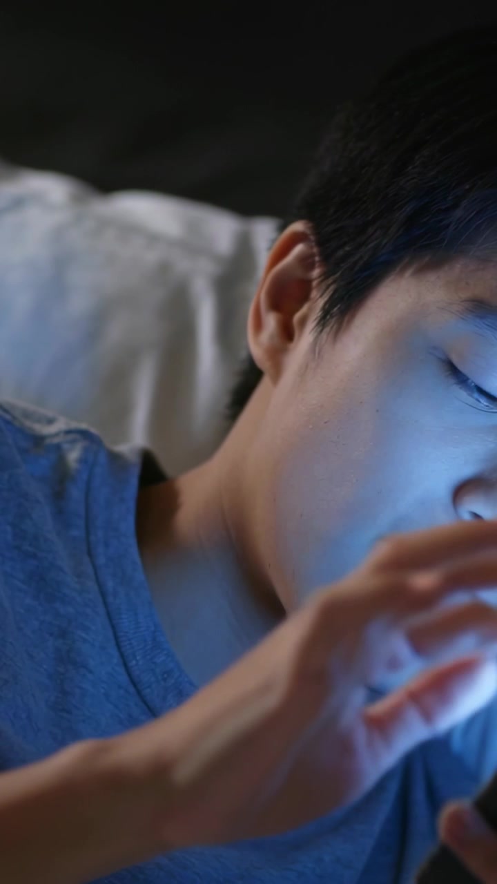 縦型動画：夜、ベッドで携帯電話を手にメッセージを確認するグレーのTシャツを着たティーンの顔に、青い光が当たっている。
