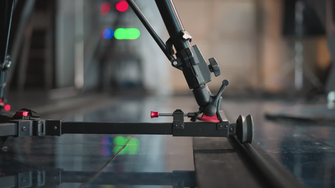Close-up of camera dolly system with wheel moving on track. Camera rig adjustment visible with focus on equipment clamps and smooth motion. Studio floor reflection adds to cinematic feel