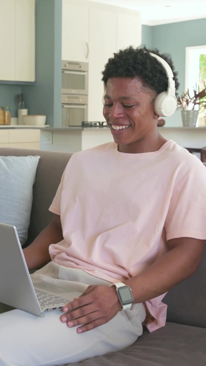 Vertical video: Hearing audio, man in pink tee smiling, typing on laptop on couch with headphones