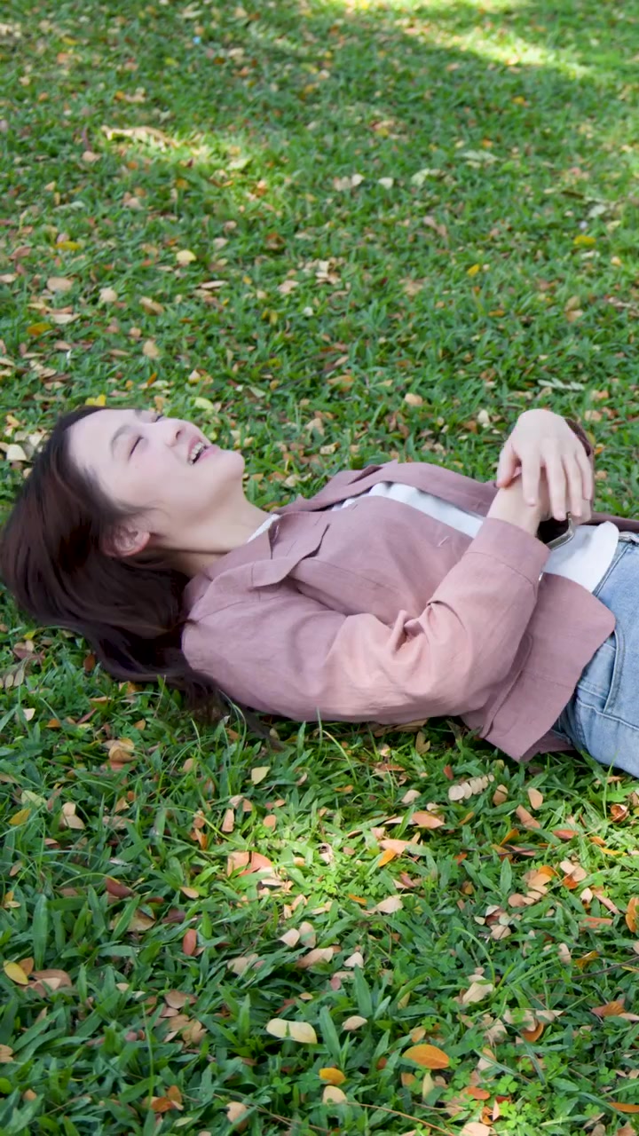 A young woman in a pink blazer sits on grass then lies down to rest