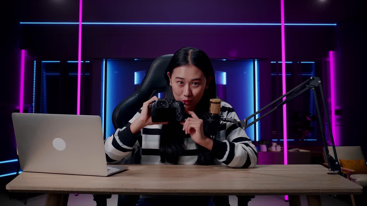 una mujer asiática sonriente con auriculares y computadora revisando la cámara en una habitación iluminada por luces de neón en retro