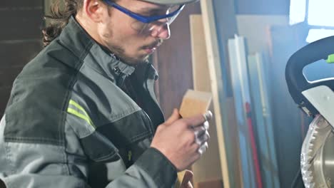 concentrated young bearded joiner in goggles and a cap checks the work piece for suitability while holding