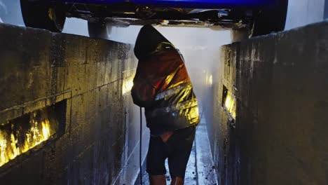 mechanic under a car with a pressurized water hose cleaning the underside of the car with a raincoat and full of dirt