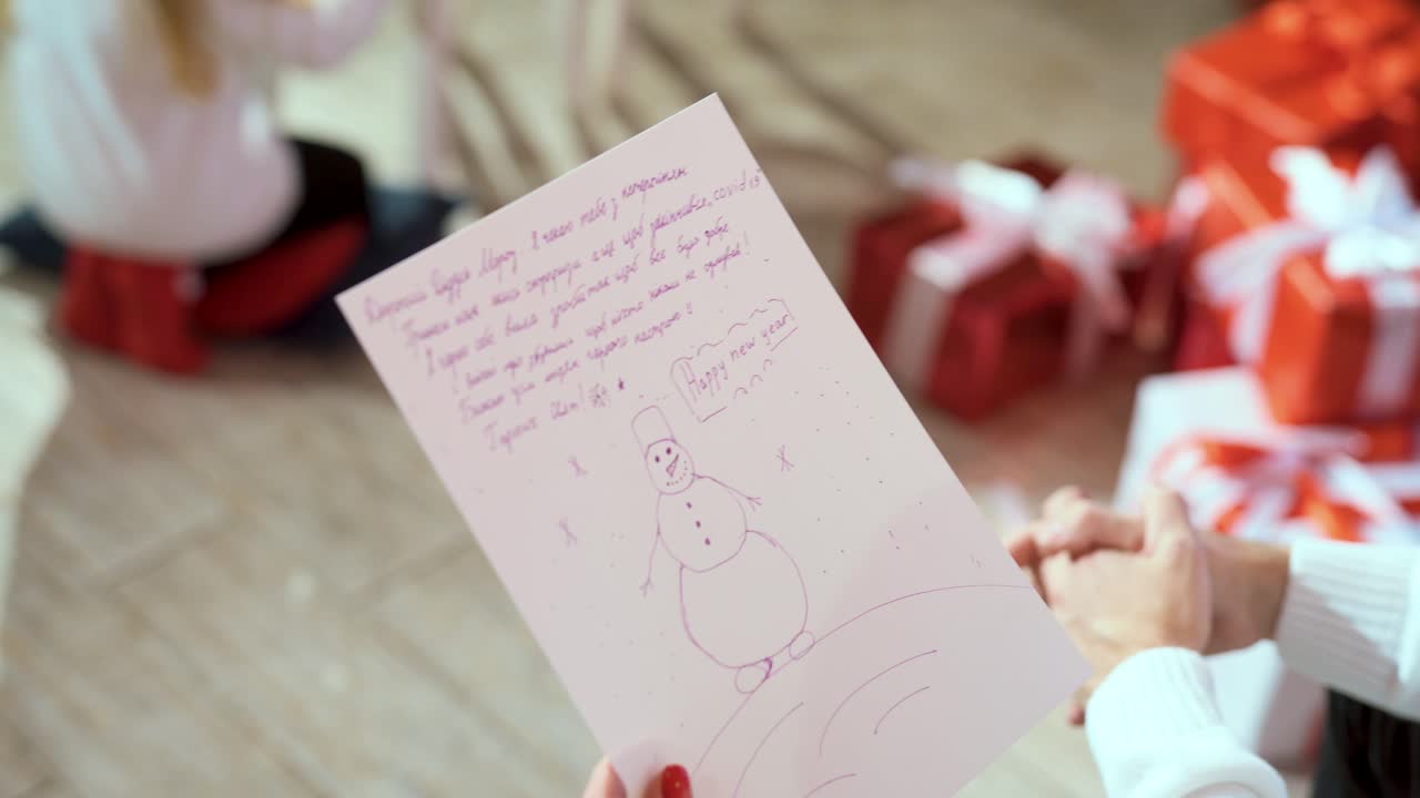 Piece of paper with a picture of a snowman and children's wishes in female hands. Letter to Santa Claus. Christmas gifts in boxes in the background