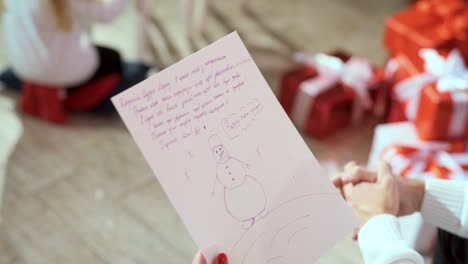 piece of paper with a picture of a snowman and children's wishes in female hands. letter to santa claus. christmas gifts in boxes in the background