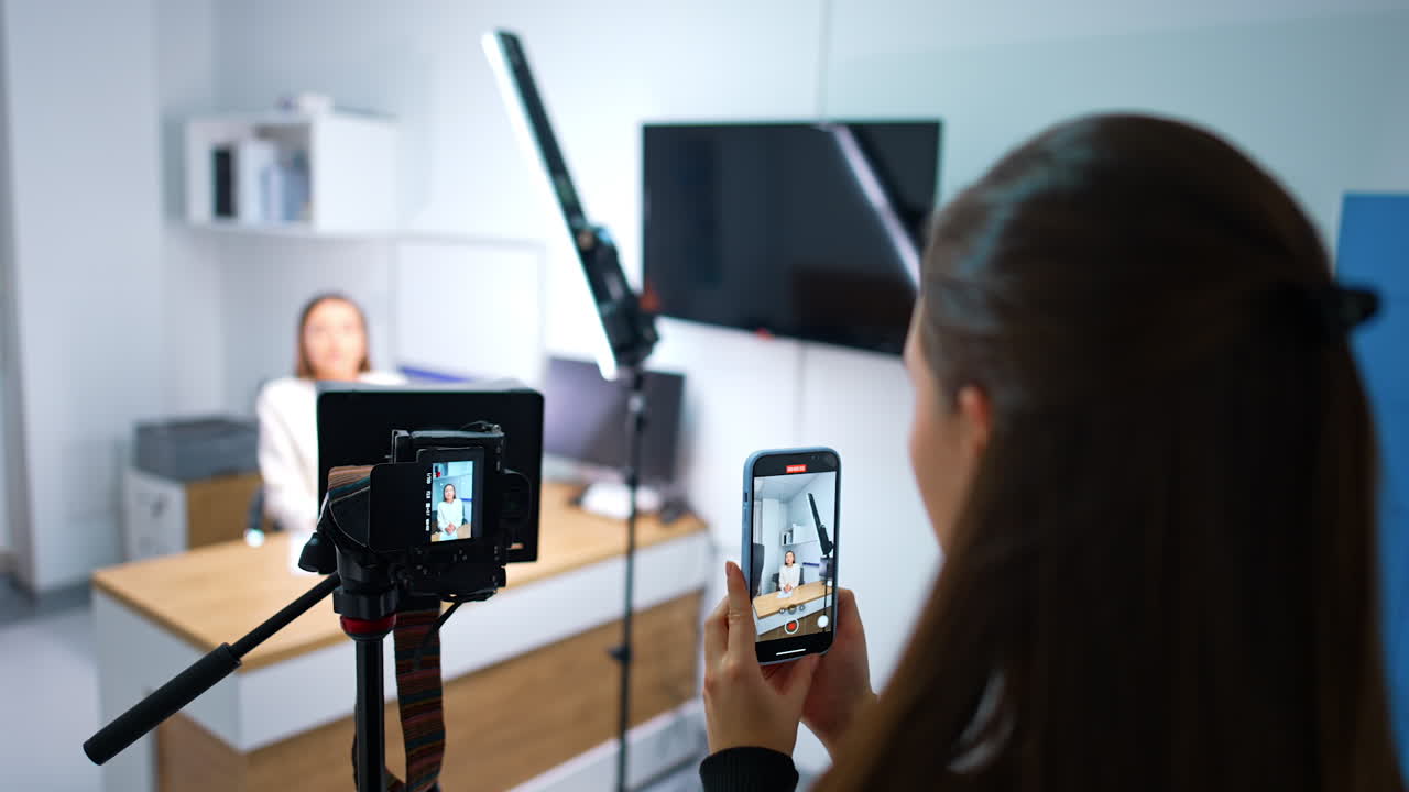 Rear view of the brunette girl taking video of a female blogger on her smartphone. Professional camera filming woman in studio. Blurred backdrop.
