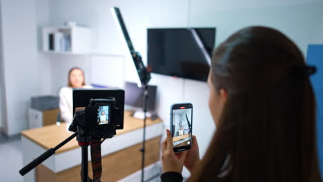 rear view of the brunette girl taking video of a female blogger on her smartphone. professional camera filming woman in studio. blurred backdrop.