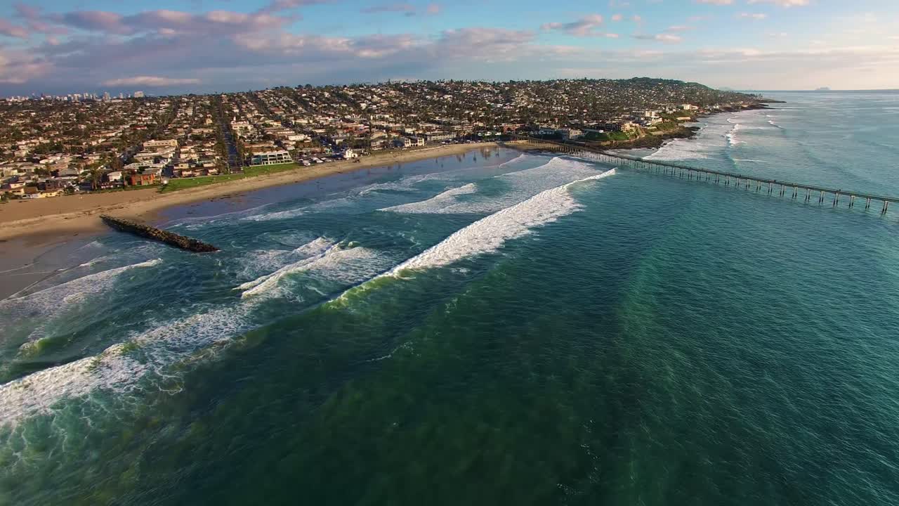 Ampia veduta aerea in 4K con drone del molo della California e della costa di una città balneare, con enormi onde blu turchese dell'Oceano Pacifico che si infrangono