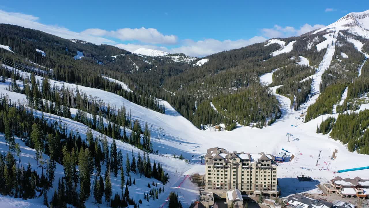 Aerial drone shot flying over Big Sky, Montana in winter, showcasing a stunning ski resort surrounded by snow-covered mountains, pine trees, and scenic alpine views in a crisp mountain atmosphere