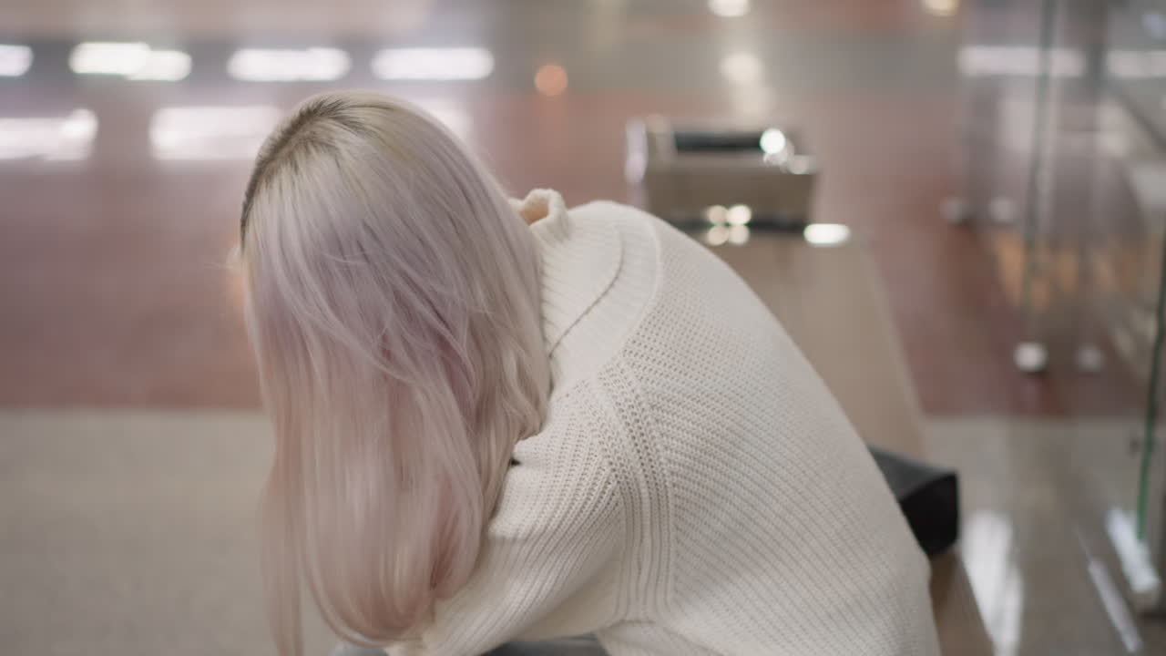 high angle view of shopper seated mall bench speaking on phone expressing thoughtfulness wearing sweater and jeans purse beside her against glass railing with blurred decorative lights background