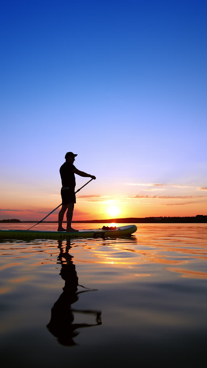 Charming sunset on the river in summer. Man on the sup board rowing with an oar appears. Low angle view. Vertical video.