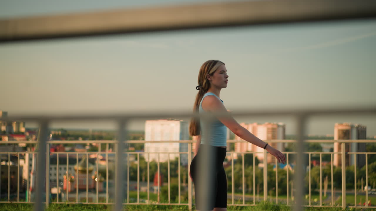 은 날 야외에서 롤러 스케이팅하는 젊은 여성, 전면에 철의 레일링, 높은 건물의 흐릿한 도시 풍경, 초록색 나무, 배경에 해가 지는 빛