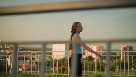 young lady roller skating outdoors on sunny day, with iron railing in foreground, blurred cityscape of tall buildings, green trees, and sunset glow in background