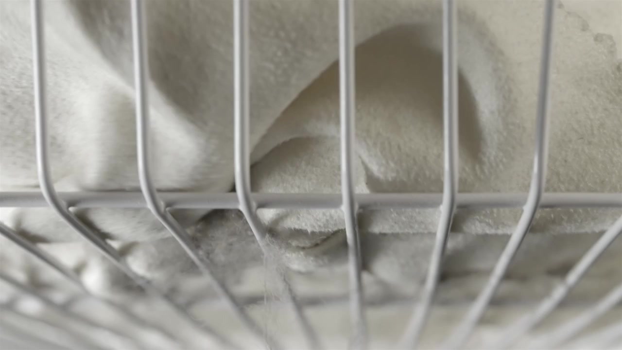 Griglia bianca di un ventilatore oscillante riempie l'inquadratura con una pala beige del ventilatore dietro.