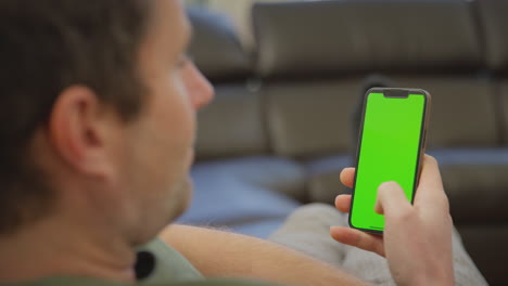 Primer-Plano-De-Un-Hombre-En-Pijama-Tumbado-En-Un-Sofá-En-Casa-Mirando-Un-Teléfono-Móvil-Con-Pantalla-Verde---Filmado-En-Cámara-Lenta