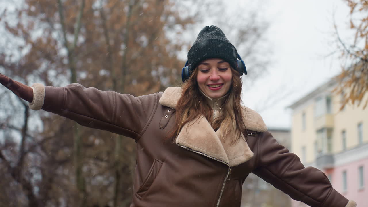 Smiling lady wearing black knit cap, brown shearling jacket with arms outstretched, walking joyfully outdoors on cool autumn day, enjoying nature and fresh air amidst vibrant autumn colors