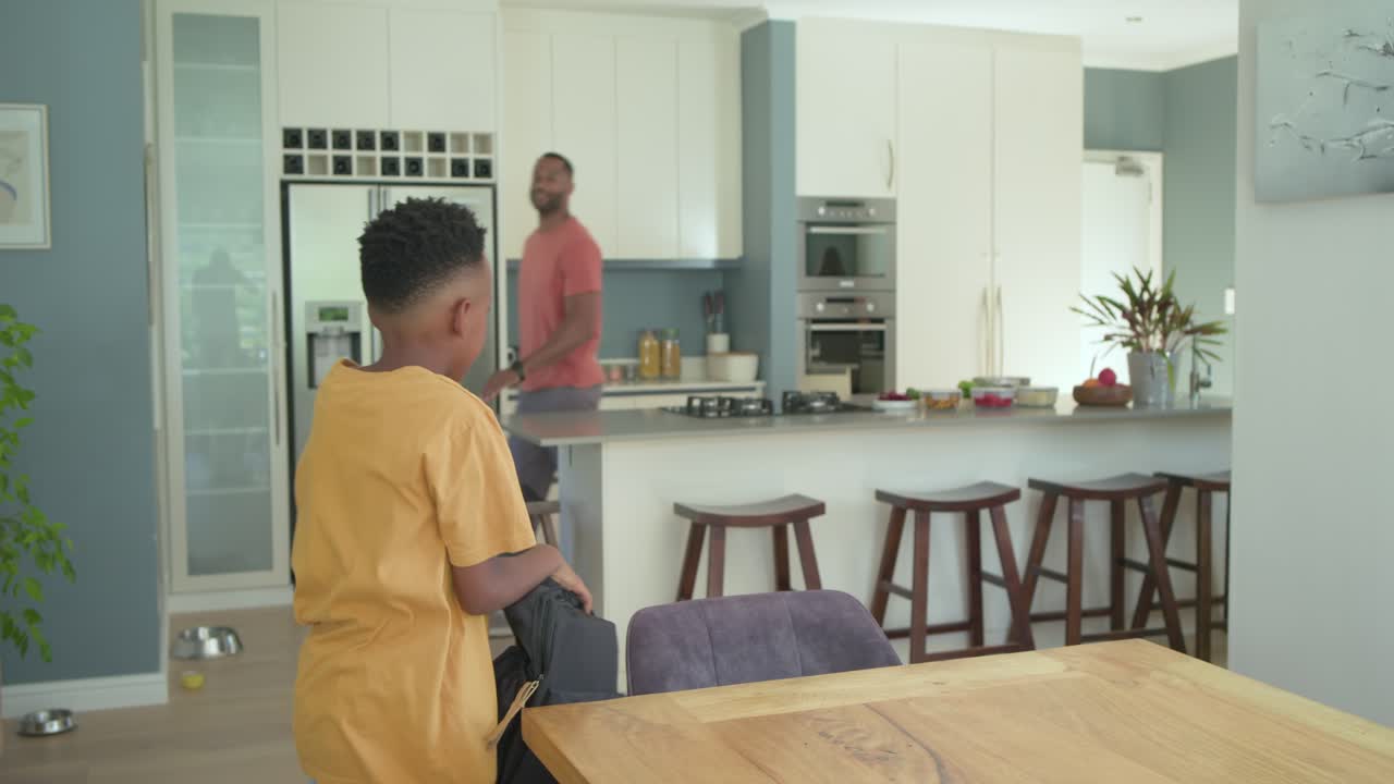 Padre e figlio afroamericani che entrano in casa con lo zaino, il padre che abbraccia e li accompagna in cucina