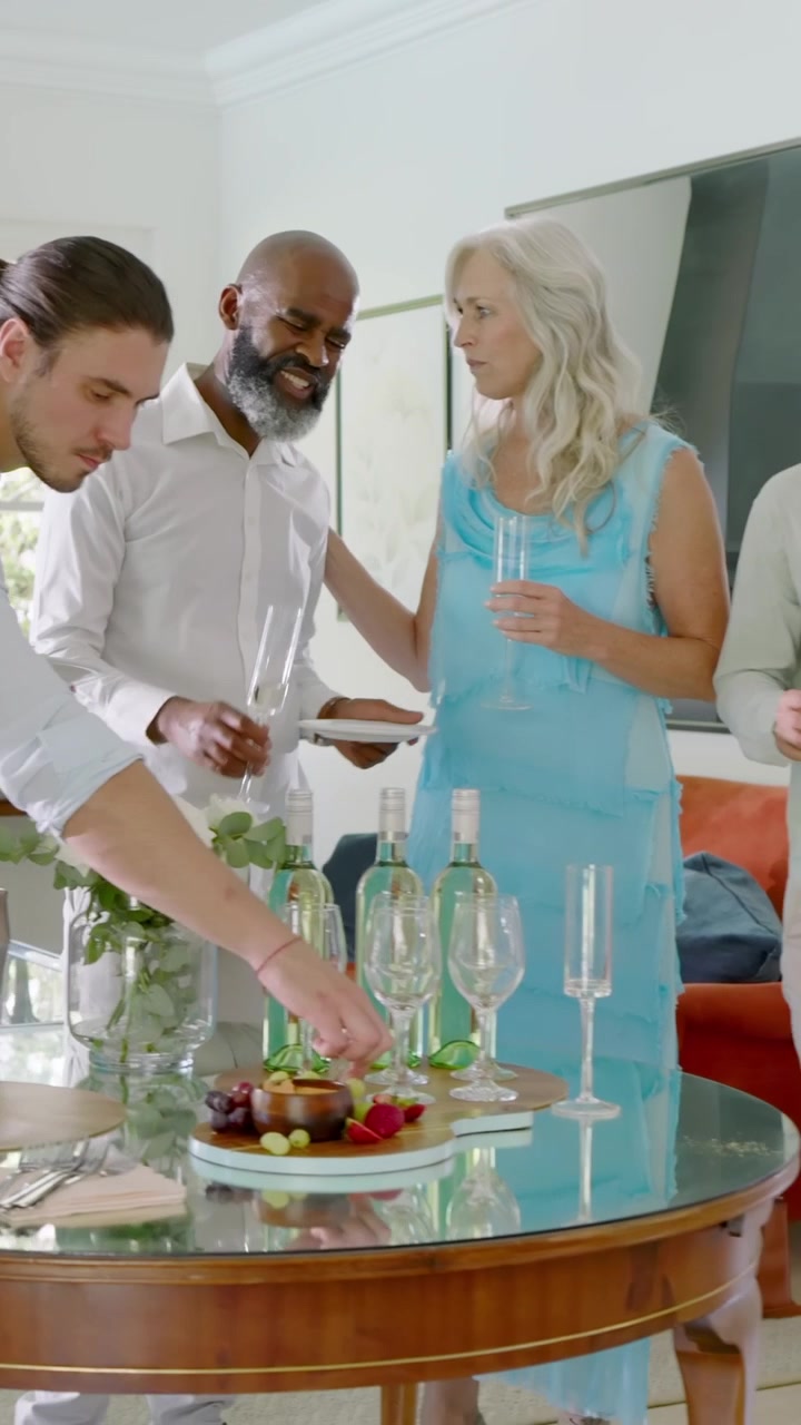 Vertical video: Diverse friends celebrating while senior woman raising flute, serving bites at home
