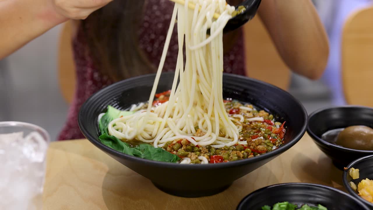 Woman uses chopsticks and spoon to mix spicy noodle soup in bright, casual dining setting