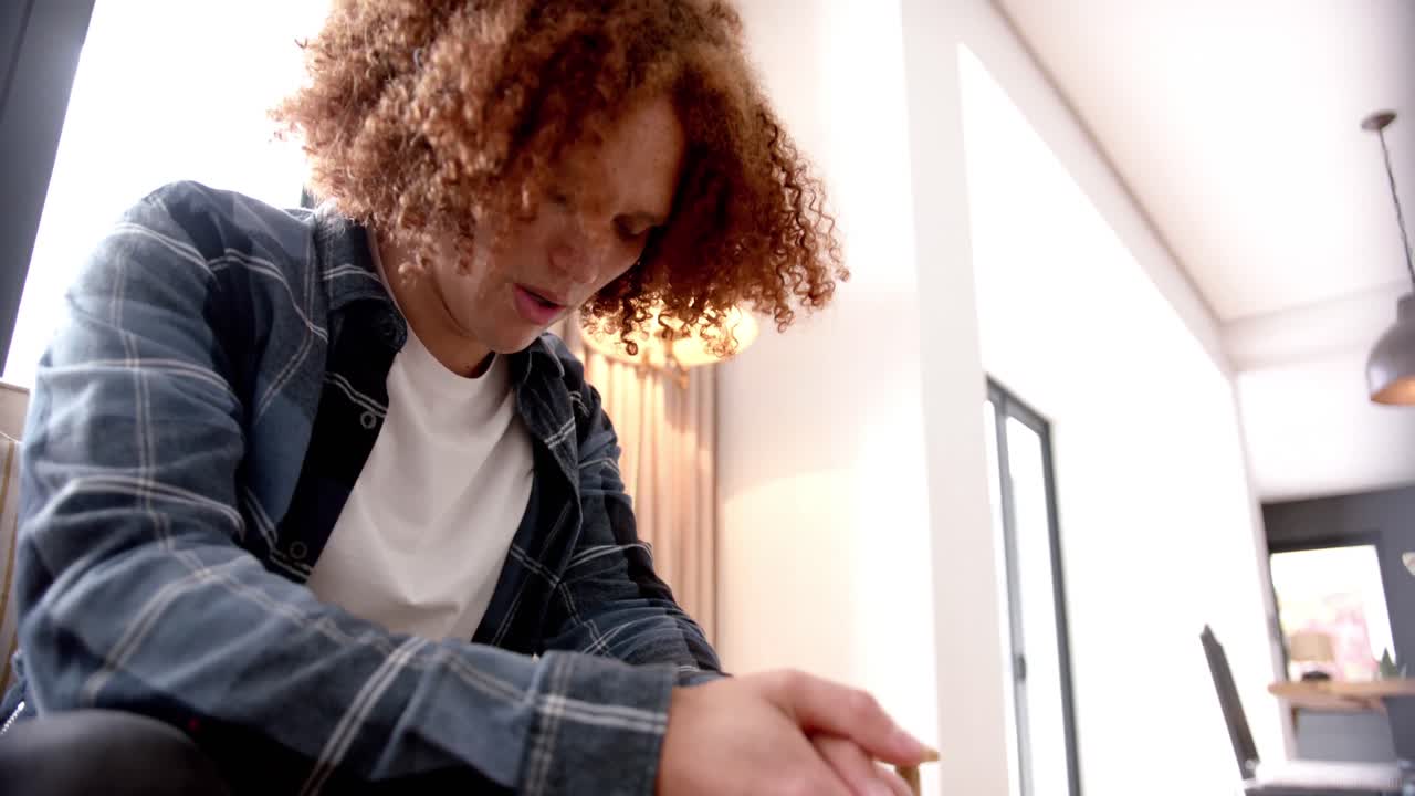 Sad biracial man with curly red hair sitting on sofa looking down in living room, slow motion
