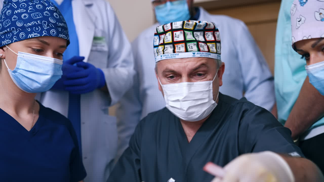 Male adult doctor in cap and mask talks to female students. Teacher shows the equipment for dental care. People in medical robes at backdrop.